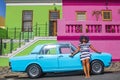 A beautiful African woman in a blue and white striped dress in front of a vintage Ford Cortina and traditional homes of Bo-Kaap, C