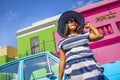 A beautiful African woman in a blue and white striped dress in front of a vintage Ford Cortina and traditional homes of Bo-Kaap, C