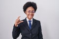 Beautiful african woman with curly hair wearing business jacket and glasses smiling and confident gesturing with hand doing small Royalty Free Stock Photo
