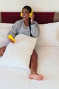 Beautiful African woman sitting on her bed in her pajamas listening to music with her cell phone and headphones Royalty Free Stock Photo