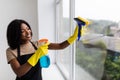 Beautiful young african woman is using a duster and a spray, looking at camera and smiling while cleaning windows in the house Royalty Free Stock Photo