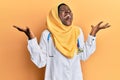 Beautiful african young woman wearing doctor uniform and hijab celebrating mad and crazy for success with arms raised and closed Royalty Free Stock Photo