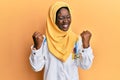 Beautiful african young woman wearing doctor uniform and hijab excited for success with arms raised and eyes closed celebrating Royalty Free Stock Photo