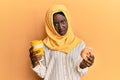 Beautiful african young woman wearing islamic hijab drinking a coffee using smartphone in shock face, looking skeptical and