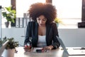 Beautiful afro business woman sending messages with her mobile phone while working with laptop in the office at home Royalty Free Stock Photo
