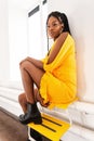 Beautiful afro woman with fashion dreadlocks in black trendy leather boots in elegant stylish yellow dress enjoys rest near white