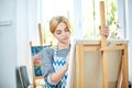 Young smiling girl paints on canvas with oil colors in own workshop. Window on the background. Art concept. Royalty Free Stock Photo