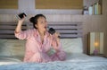 Beautiful Asian American teenager girl singing karaoke song excited at home bedroom holding mobile phone playing on bed excited Royalty Free Stock Photo