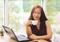 Beautiful Asian business woman sitting alone in the coffee shop with computer laptop on the table holding cup of coffee smiling Royalty Free Stock Photo