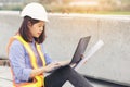 Beautiful Asian female engineer in white safety hard hat using laptop computer notebook doing job at construction site outside Royalty Free Stock Photo