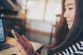 Asian girl with smiley face using smart phone with laptop on wooden table in modern cafe Royalty Free Stock Photo