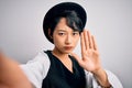 Beautiful asian girl wearing casual hat making selfie by the camera over white background with open hand doing stop sign with Royalty Free Stock Photo