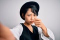 Beautiful asian girl wearing casual hat making selfie by the camera over white background pointing with finger to the camera and Royalty Free Stock Photo