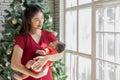 Beautiful Asian mom woman wear red cloth holding newborn baby in arms standing looking outside windows at cozy home.Happy infant Royalty Free Stock Photo