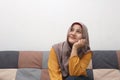 Beautiful Asian muslim woman thinking something good while sitting on sofa, girl with contemplation expression, looking up and
