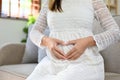 Pregnant woman touching her belly with love, making a heart sign with hands while sitting on sofa Royalty Free Stock Photo