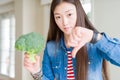 Beautiful Asian woman eating green fresh broccoli with angry face, negative sign showing dislike with thumbs down, rejection Royalty Free Stock Photo