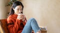 Beautiful Asian woman enjoy a cup of hot cocoa or chocolate with marshmallow while sit on chair in living room at home. Korea girl Royalty Free Stock Photo