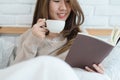 Beautiful asian woman enjoying warm coffee and reading book on bed in her bedroom.