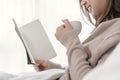 Beautiful asian woman enjoying warm coffee and reading book on bed in her bedroom. Royalty Free Stock Photo