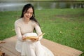 A beautiful Asian woman enjoys reading a book while picnicking in a green park on the weekend alone