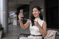 A beautiful Asian woman is having a video call on her smartphone while sitting on a bench outdoors Royalty Free Stock Photo