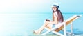 Beautiful Asian woman holds glasses and towels, wears a hat yellow sandals. Sitting smiling on a chair at the beach, blue sky as a Royalty Free Stock Photo