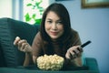 Beautiful Asian woman laying on sofa in relax and happy holding remote control in hand with bowl of popcorn in home living room Royalty Free Stock Photo