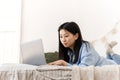 A beautiful Asian woman is lying on the bed and working using a laptop from home. A young woman works at home in