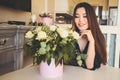 Beautiful Asian woman sits by the table in living room bright interior admires white roses flowers bouquet. Smiling Royalty Free Stock Photo
