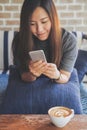 A beautiful Asian woman with smiley face using and looking at smart phone sitting on sofa with white latte coffee cup Royalty Free Stock Photo