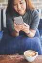Asian woman with smiley face using and looking at smart phone sitting on sofa with white latte coffee cup on wooden table