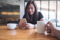 A beautiful Asian woman with smiley face using smart phone with a man hand holding white coffee cup Royalty Free Stock Photo