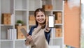 Beautiful Asian woman smiling holding clipboard and hands showing smartphone mockup of blank screen in stock room Royalty Free Stock Photo