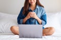 A beautiful asian woman using laptop computer for video call while sitting and drinking coffee on a bed at home Royalty Free Stock Photo