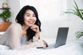 Beautiful Asian woman using laptop lying down in bed smiling to the camera, work from home, shopping online concept Royalty Free Stock Photo