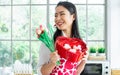 Beautiful Asian woman wearing apron with heart pattern, smiling with happiness, holding flowers, gift box of cookies, standing in Royalty Free Stock Photo