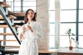 Beautiful Asian woman in white robe holding cup of coffee, standing against the backdrop of staircases Royalty Free Stock Photo