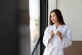 Beautiful Asian woman in white robe holding cup of coffee, standing near the window. Royalty Free Stock Photo