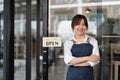 Beautiful asian young barista woman in apron holding tablet and standing in front of the door of cafe with open sign Royalty Free Stock Photo