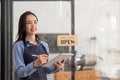 Beautiful asian young barista woman in apron holding tablet and standing in front of the door of cafe with open sign Royalty Free Stock Photo