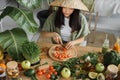 Beautiful asian young woman in traditional conical hat is slicing cherry tomatoes, sitting at table Royalty Free Stock Photo