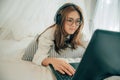 Beautiful asian young woman wearing glasses and headphones listening music or watching movie with laptop on the white bed at home. Royalty Free Stock Photo