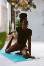 Beautiful and athletic afro american woman sitting and pulling bent rear leg, doing yoga on gymnastic mat in decorated