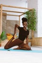 Beautiful and athletic afro american woman sitting with smile and pulling bent rear leg, doing yoga on gym mat in