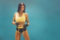 Beautiful athletic girl in yellow top and black shorts standing at blue wall and holding measuring strap around waist Royalty Free Stock Photo