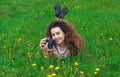 Beautiful, attractive girl-photographer with curly hair holds a camera and lying on the grass with blooming dandelions Royalty Free Stock Photo