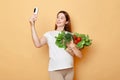 Beautiful attractive pregnant woman holding fresh vegetables posing isolated over beige background making selfie on cell phone Royalty Free Stock Photo