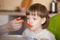 Beautiful baby eats porridge from mom`s hand. The mother feeds the son with a spoon. Boy with long blond hair and big blue eyes p Royalty Free Stock Photo