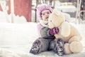 Beautiful baby embracing toy plush bear sitting on snow in park in cold sunny winter day during winter holidays